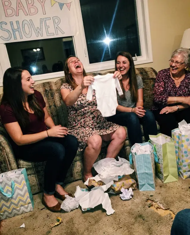 A mum-to-be laughing on the couch as she holds up a tiny onesie